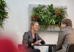 The lady sitting on the table with man and behind them there is  a plant hanging on the wall.